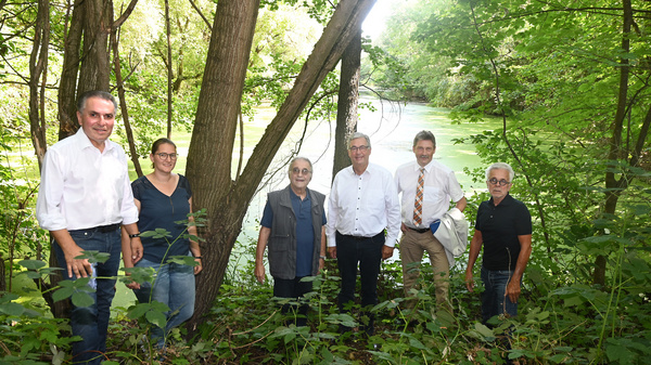 Un gruppo di sei persone si trova in una zona boschiva vicino a un lago. Tre uomini e tre donne sorridono, vestiti in modo casual. La vegetazione è verde e l'acqua del lago è visibile sullo sfondo, creando un'atmosfera naturale e tranquilla.