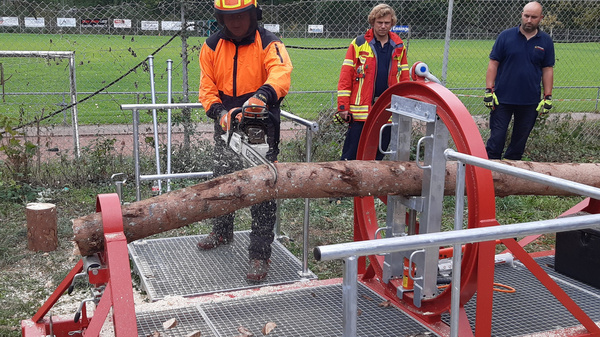 A person in safety gear operates a chainsaw on a log positioned on a cutting machine, while two individuals observe nearby. The setting is outdoors, with a grassy area and trees in the background.