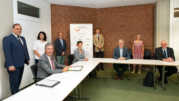 A formal meeting setting is depicted, with a group of eight people gathered around a long table. A man in a suit is signing a document at the table's forefront. The background features a banner and a brick wall. Participants appear engaged and attentive.
