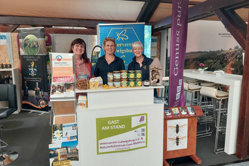 Drei lächelnde Frauen stehen an einem Messestand. Sie präsentieren Naturprodukte und regionale Spezialitäten. Im Hintergrund sind verschiedene Informationsmaterialien und Banner sichtbar, die auf den Verein und das Naturerlebniszentrum hinweisen. Der Stand ist hell und einladend gestaltet. (Text KI generiert)