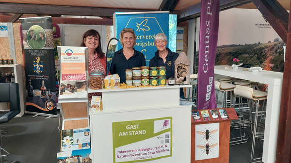 Drei lächelnde Frauen stehen an einem Messestand. Sie präsentieren Naturprodukte und regionale Spezialitäten. Im Hintergrund sind verschiedene Informationsmaterialien und Banner sichtbar, die auf den Verein und das Naturerlebniszentrum hinweisen. Der Stand ist hell und einladend gestaltet. (Text KI generiert)