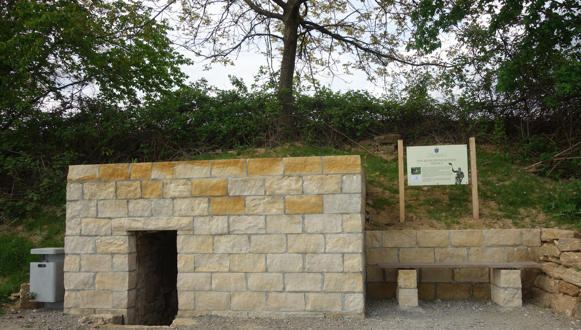 Ein kleines gemauerte Gebäude mit einer offenen Tür steht in einer natürlichen Umgebung. Daneben befindet sich eine Holztafel mit Informationen. Im Vordergrund sind ein Tisch und eine Bank aus Stein. Grüne Büsche und ein Baum sind im Hintergrund sichtbar.