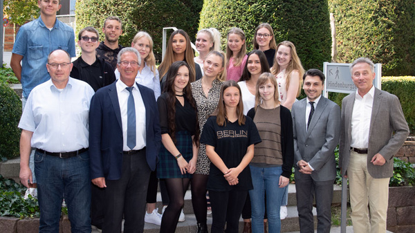 A diverse group of 20 people stands together outdoors in front of lush, rounded hedges. They are dressed in various formal and casual attire, smiling and posing for the photo, suggesting a celebratory or formal occasion.
