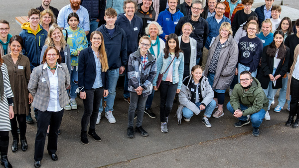 Eine große Gruppe von etwa 50 Menschen steht im Freien, umgeben von Bäumen und Tischbänken. Die Personen lächeln und posieren, als wäre es ein Gruppenfoto, wohl aus einer Veranstaltung oder einem Treffen. Alle tragen unterschiedliche Kleidungsstile.