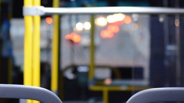 Interior of an empty bus featuring yellow support poles, blue upholstered seats, and dim lighting. The view focuses on the seats with a blurry background of the bus's interior and illuminated signage, suggesting a quiet, waiting atmosphere.
