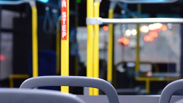 Interior of an empty bus featuring yellow support poles, blue upholstered seats, and dim lighting. The view focuses on the seats with a blurry background of the bus's interior and illuminated signage, suggesting a quiet, waiting atmosphere.