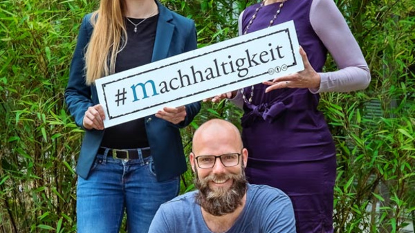 Trois personnes posent devant un décor de verdure. Deux femmes tiennent une pancarte avec le mot-clé « #Machhaltigkeit » (durabilité en allemand) et un homme est accroupi à leurs côtés. L'image évoque un engagement pour des pratiques durables.
