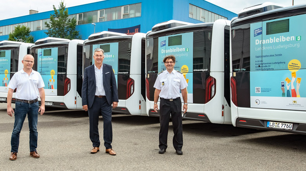 Trois personnes en tenue professionnelle se tiennent devant une rangée de bus affichant des panneaux d'information sur un projet ou une initiative. L'image met en avant l'engagement envers le transport public et la sensibilisation communautaire.