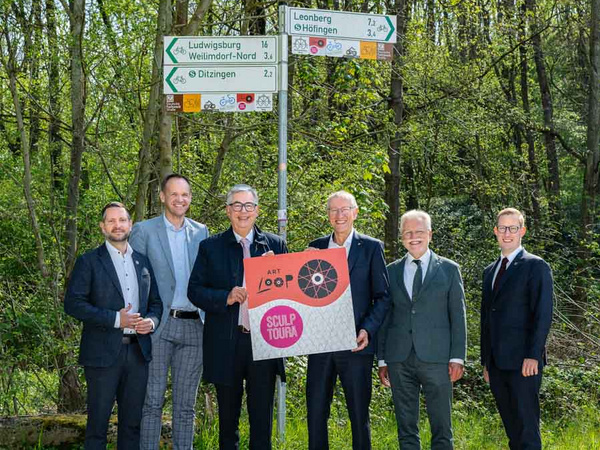 20.04.2026 - Eröffnung der Verbindungsstrecke Art Loop & Sculptoura mit dem Böblinger Landrat Roland Bernhard Sechs Männer stehen an einem Wegweiser im Freien. Sie halten ein Schild mit der Aufschrift "ART LOOP SCULPTURA" in den Händen. Im Hintergrund sind Bäume und die Wegweiser mit verschiedenen Richtungshinweisen sichtbar. Es ist ein sonniger Tag.