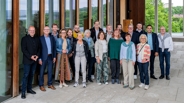 Eine Gruppe von etwa 20 Personen steht im Freien vor einem Gebäude mit großen Fenstern. Die Teilnehmer sind unterschiedlich gekleidet und lächeln. Die Umgebung ist grün und die Atmosphäre wirkt freundlich und einladend.