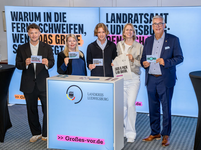 Eine Gruppe von fünf Personen steht vor einem beleuchteten Stand mit Informationen über den Landkreis Ludwigsburg. Sie halten Schilder mit verschiedenen Botschaften in den Händen und lächeln in die Kamera. Im Hintergrund sind auffällige Banner zu sehen. (Text KI generiert)