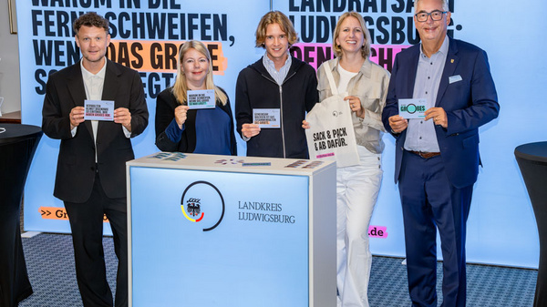 Eine Gruppe von fünf Personen steht vor einem beleuchteten Stand mit Informationen über den Landkreis Ludwigsburg. Sie halten Schilder mit verschiedenen Botschaften in den Händen und lächeln in die Kamera. Im Hintergrund sind auffällige Banner zu sehen. (Text KI generiert)