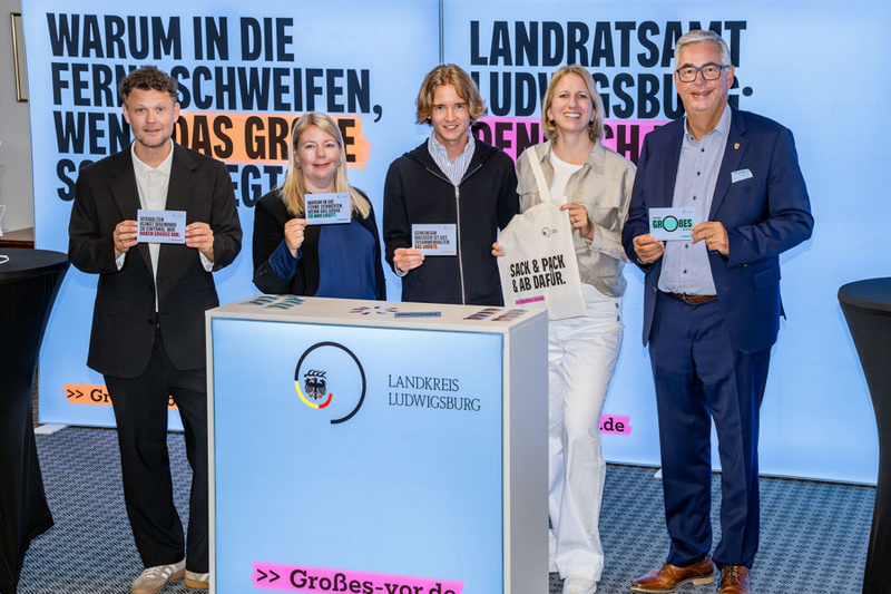 Eine Gruppe von fünf Personen steht vor einem beleuchteten Stand mit Informationen über den Landkreis Ludwigsburg. Sie halten Schilder mit verschiedenen Botschaften in den Händen und lächeln in die Kamera. Im Hintergrund sind auffällige Banner zu sehen. (Text KI generiert)