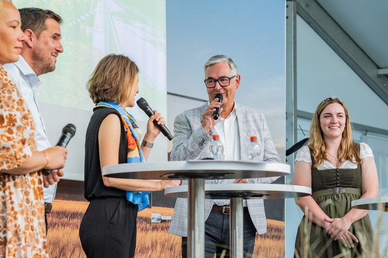 Eine Gruppe von vier Personen steht an einem Tisch auf einer Bühne. Sie halten Mikrofone und wirken in ein Gespräch vertieft. Die Hintergrundgrafik zeigt eine Landschaft. Die Personen sind unterschiedlich gekleidet, darunter traditionelle Trachten. (Text KI generiert)