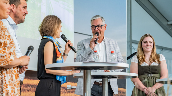 Eine Gruppe von vier Personen steht an einem Tisch auf einer Bühne. Sie halten Mikrofone und wirken in ein Gespräch vertieft. Die Hintergrundgrafik zeigt eine Landschaft. Die Personen sind unterschiedlich gekleidet, darunter traditionelle Trachten. (Text KI generiert)