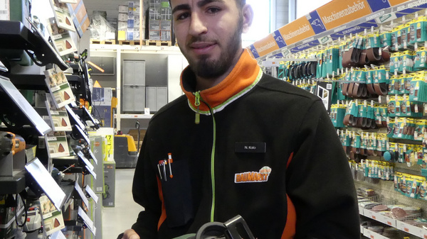 A young man in a black and orange work uniform holds a power tool in a hardware store. The shelves are filled with various tools and equipment, indicating a well-stocked DIY section. The setting suggests a focus on home improvement and construction supplies.