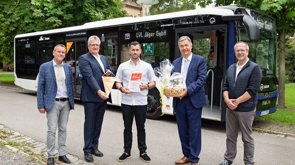 Cinq hommes se tiennent devant un bus. Ils tiennent des cadeaux et des brochures. L'ambiance est conviviale, probablement pour une cérémonie ou un événement spécial. Le bus est identifié comme appartenant à une entreprise de transport.