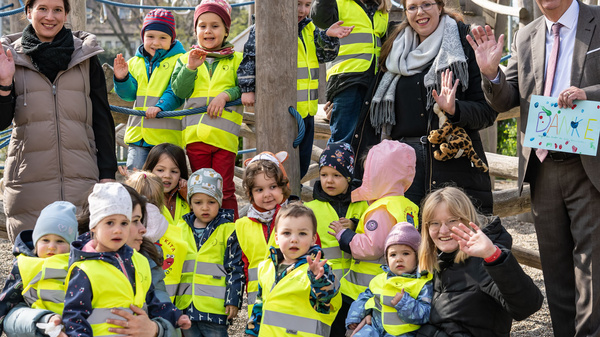 Eine Gruppe von Kindern in leuchtenden Sicherheitswesten steht auf einem Spielplatz. Sie sind umgeben von Erwachsenen, die lächeln und die Hände zum Winken heben. Die Szene vermittelt Freude und Gemeinschaft. Im Hintergrund sind Spielgeräte sichtbar.