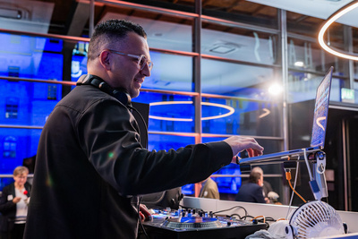 Ein DJ steht an einem Mischpult in einem modernen Veranstaltungsort. Er nutzt Kopfhörer und konzentriert sich auf seinen Laptop, während er Musik auflegt. Blaues Licht beleuchtet den Raum, und im Hintergrund sind weitere Personen zu sehen, die sich unterhalten.