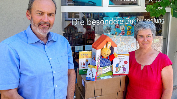 A man and a woman stand in front of a bookstore window filled with books and a colorful display. The woman, wearing a red dress, holds a book, while a puppet resembling a character from children's literature is positioned beside them. Boxes are stacked behind them.
