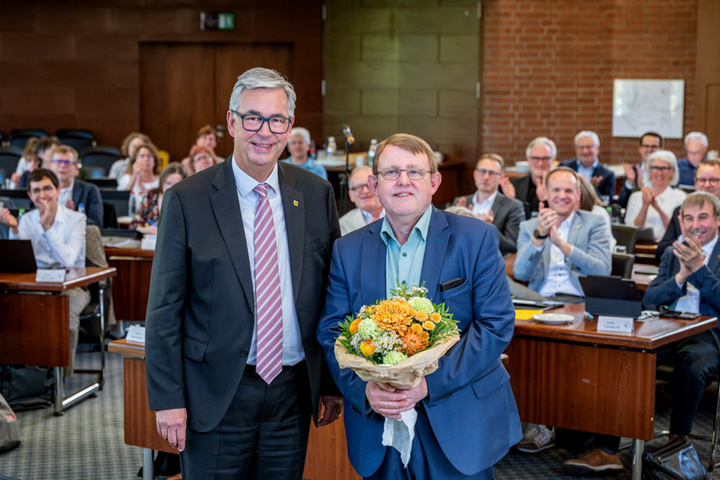 Zwei Männer stehen in einem Konferenzraum. Der Links trägt einen Anzug mit Krawatte und lächelt, während der Rechts einen Strauß Blumen hält und ebenfalls lächelt. Im Hintergrund applaudiert eine Gruppe von Menschen. Die Szene vermittelt Anerkennung und Freude.