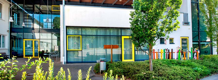 Header Bild: Das Bild zeigt das modern gestaltete Gebäude einer Einrichtung, mit großen Fenstern und einem charakteristischen Holzdach. Auf der linken Seite sind bunte Holzzäune und grüne Pflanzen zu sehen, die das freundliche Ambiente unterstreichen.