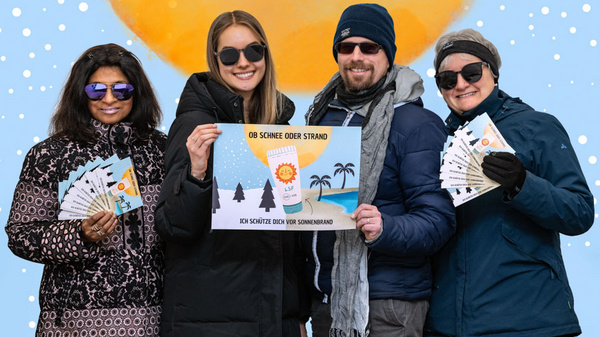 Vier Personen stehen zusammen und halten ein Plakat, auf dem steht: „Ob Schnee oder Strand – ich schütze dich vor Sonnenbrand.“ Sie tragen Sonnenbrillen und lächeln. Im Hintergrund ist eine große Sonne und Illustrationen von Palmen und Schneebäumen sichtbar.