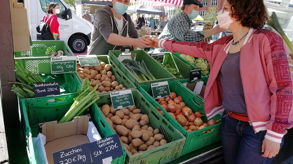 A market vendor hands a customer fresh produce, including zucchini, potatoes, and apples, displayed in green baskets. Both individuals are wearing face masks. The scene captures a vibrant, lively atmosphere of a farmer's market.