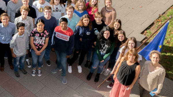 A diverse group of approximately 50 students stands together in a spacious outdoor area, smiling and looking at the camera. They are dressed casually, with some holding flags. The scene is well-lit, showcasing the camaraderie among the group.