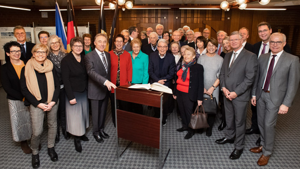 A diverse group of over thirty people, including men and women of various ages, gathers around a podium with a large book on it. Some individuals are smiling, dressed in formal and semi-formal attire, suggesting a celebratory or significant event. Flags are visible in the background.