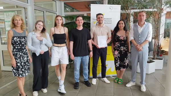 Eine Gruppe von sechs Personen steht in einem hellen Raum. Sie lächeln und posieren zusammen, während einer von ihnen ein Dokument in der Hand hält. Im Hintergrund ist ein Banner der "Robert-Franck-Schule" sichtbar. Pflanzen und große Fenster verleihen dem Raum eine freundliche Atmosphäre.