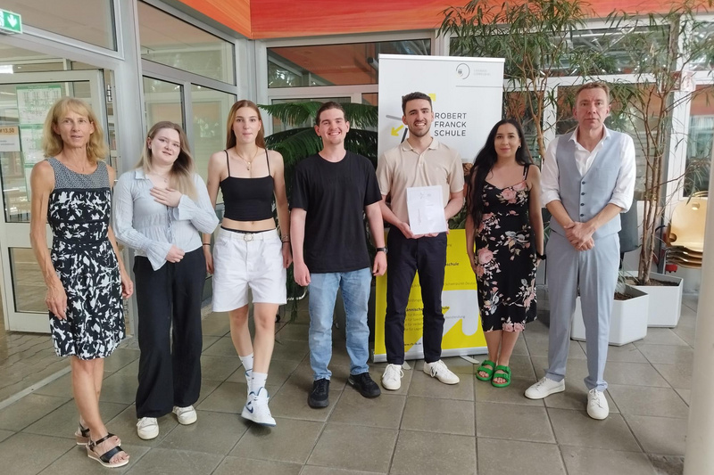 Eine Gruppe von sechs Personen steht in einem hellen Raum. Sie lächeln und posieren zusammen, während einer von ihnen ein Dokument in der Hand hält. Im Hintergrund ist ein Banner der "Robert-Franck-Schule" sichtbar. Pflanzen und große Fenster verleihen dem Raum eine freundliche Atmosphäre.