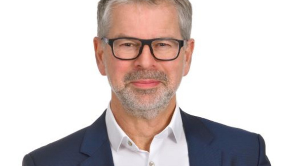 Porträt eines Mannes mittleren Alters mit kurzen grauen Haaren und Brille. Er trägt einen blauen Anzug und ein weißes Hemd, wirkt freundlich und selbstbewusst. Der Hintergrund ist neutral und hell.