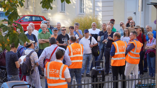 Eine Gruppe von Menschen versammelt sich im Freien, während Personen in orangefarbener Kleidung, die auf eine Veranstaltung hinweisen, um sie herumstehen. Die Umgebung wirkt lebhaft und informell, mit Bäumen im Hintergrund und Fahrrädern in der Nähe.