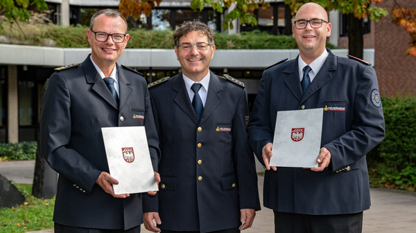 Drei Männer in Uniform stehen lächelnd im Freien. Sie halten jeweils ein Dokument in der Hand. Die Szene vermittelt einen feierlichen Anlass, wahrscheinlich eine Auszeichnung oder Ehrung. Im Hintergrund sind Bäume und ein Gebäude sichtbar.