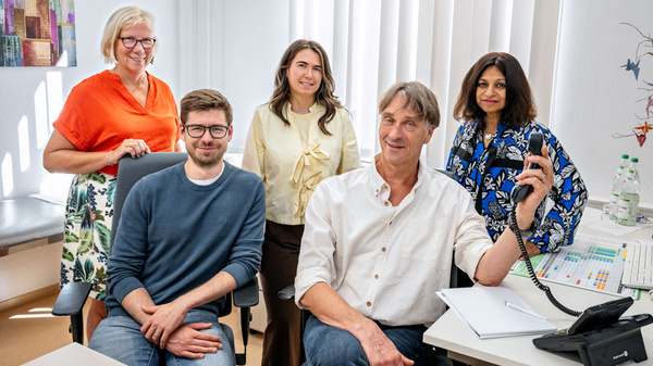 Fünf Personen in einem Büroraum, lächelnd und in lockerer Atmosphäre. Ein Mann hält ein Telefon in der Hand, während die anderen um ihn herum stehen. Die Gruppe vermittelt ein Gefühl von Zusammenarbeit und Professionalität. Im Hintergrund sind Fenster mit Jalousien und ein Schreibtisch sichtbar.