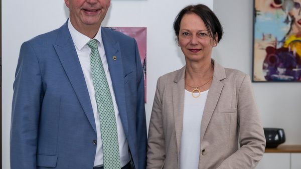 Zwei Personen stehen in einem Büro. Der Mann trägt einen hellen Anzug mit einer grünen Krawatte und lächelt. Die Frau steht neben ihm in einem hellen Blazer und hält eine Hand in der Tasche. Im Hintergrund sind stimmungsvolle Bilder an der Wand.