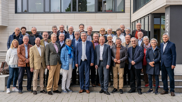Eine Gruppe von etwa 40 Personen steht lächelnd vor einem Gebäude. Die Teilnehmer sind überwiegend älteren Alters und tragen verschiedene formelle und legere Kleidung. Sie stehen in mehreren Reihen und wirken freundlich und einladend.