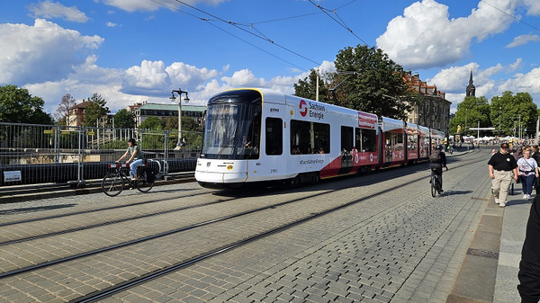 Eine Straßenbahn fährt auf Schienen entlang einer gepflasterten Straße. Einige Radfahrer und Fußgänger sind in der Nähe. Der Himmel ist blau mit einigen Wolken, und im Hintergrund sind Bäume und Gebäude sichtbar.