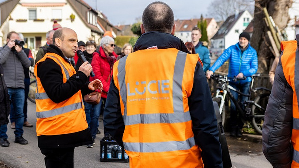 Zwei Männer in orangefarbenen Sicherheitswesten stehen auf einer Straße. Einer der Männer spricht, während eine Gruppe von Menschen im Hintergrund lauscht. Die Szene vermittelt eine Atmosphäre von Engagement und öffentlichem Interesse, möglicherweise im Rahmen einer Veranstaltung oder Diskussion.