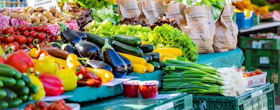 Header Bild: Ein Märkten mit frischem Obst und Gemüse, darunter Tomaten, Zucchini, Auberginen und verschiedene Salate. Im Hintergrund sind Einkaufstüten aus Papier zu sehen. Bunte Markisen schützen die Waren vor Sonne.