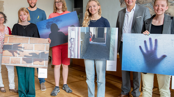 Eine Gruppe von sieben Personen steht in einem Raum und hält verschiedene große Fotos in den Händen. Die Bilder zeigen unterschiedliche Motive, während im Hintergrund eine Wand mit einem historischen Wandgemälde zu sehen ist.