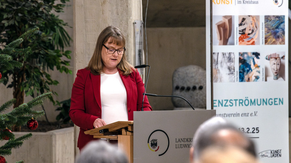 Eine Frau in einem roten Blazer steht am Rednerpult und spricht zu einem Publikum. Im Hintergrund ist ein festlich geschmückter Weihnachtsbaum sichtbar, sowie ein Plakat mit Informationen zur Kunstausstellung „ENZSTRÖMUNGEN“. Der Raum hat eine ruhige, feierliche Atmosphäre.
