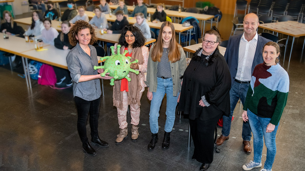 Un groupe de six personnes se tient debout dans une salle avec des tables et des élèves assis en arrière-plan. L'une des femmes tient une marionnette en forme de virus. Elles sourient, représentant un message de sensibilisation ou d’éducation sanitaire.