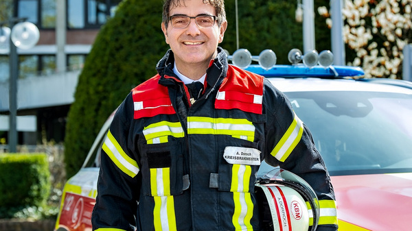 Ein Feuerwehrmann steht lächelnd vor einem Einsatzfahrzeug. Er trägt eine Feuerwehruniform mit reflektierenden Streifen und hält einen Feuerwehrhelm in der Hand. Im Hintergrund sind grüne Sträucher und ein Gebäude zu sehen.