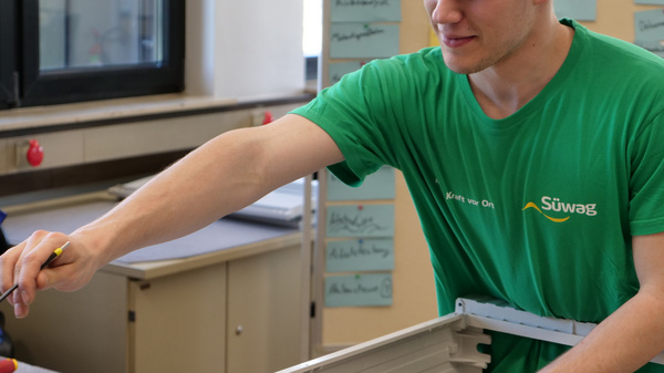 Ein junger Mann in einem grünen T-Shirt arbeitet konzentriert an einem elektronischen Gerät. Er hält Werkzeug in der Hand und ist dabei, Kabel zu verbinden oder zu installieren. Im Hintergrund sind Arbeitsmaterialien und Notizen sichtbar.