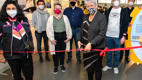 Eine Gruppe von Personen steht in einem Ausstellungsraum, um eine Eröffnung zu feiern. Eine Frau schneidet ein rotes Band durch, während andere Personen, die Masken tragen, zuschauen. Im Hintergrund sind informative Displays und eine große, gelbe Installation zu sehen.