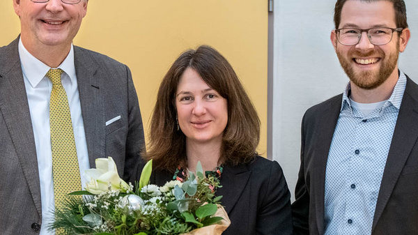 Three people stand together, smiling. The woman in the center holds a bouquet of flowers, while the two men on either side of her are dressed in formal attire. The background features a yellow wall and a neutral-colored space, suggesting a celebratory event.