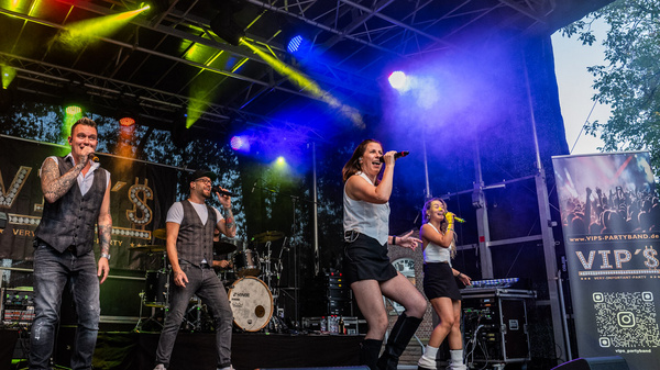 Eine Band führt auf einer Bühne ein Live-Konzert auf. Mitglieder der Band spielen Instrumente und singen, während bunte Lichter die Atmosphäre beleuchten. Die Stimmung ist energetisch und lebhaft.