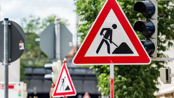 Ein Baustellenschild mit einem Symbol, das einen Arbeiter mit Schaufel darstellt, ist im Vordergrund zu sehen. Dahinter sind mehrere Verkehrsschilder zu erkennen. Das Bild zeigt eine Stadtlandschaft mit Bäumen im Hintergrund. (Text KI generiert)